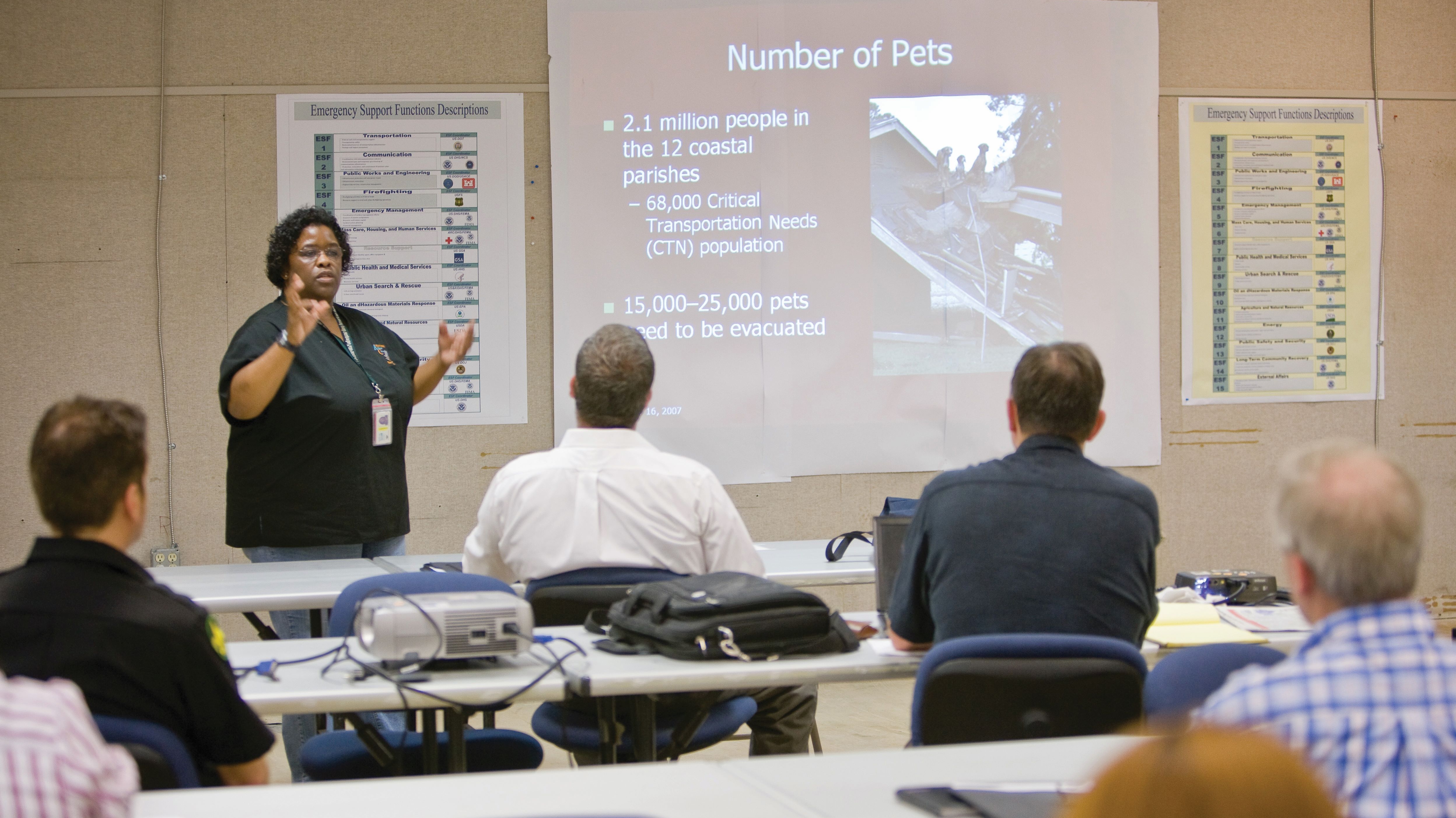 Woman speaker standing next to a large screen presenting to a group of people