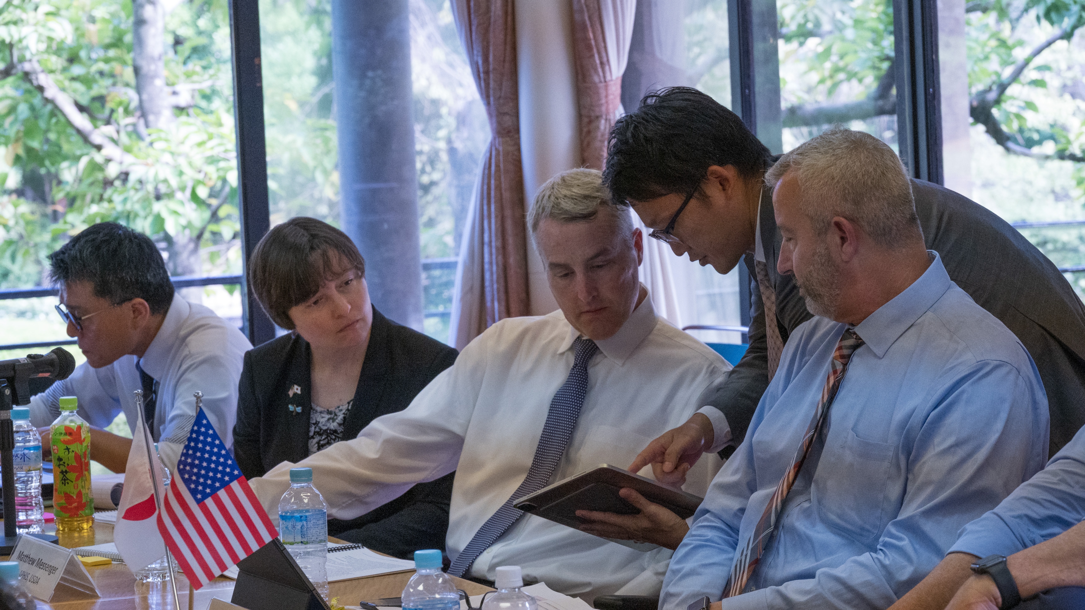 Group of people sitting a table while another person leans in to show them something on a tablet.